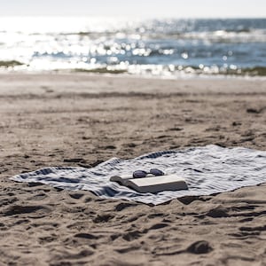 May include: A white book and sunglasses on a blue and white striped beach towel on a sandy beach.