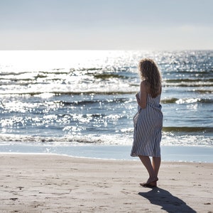 May include: A woman in a striped dress stands on a sandy beach, looking out at the ocean. The water is blue and sparkling in the sunlight.