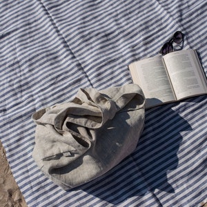 May include: A white linen tote bag with a blue and white striped beach blanket, a book, and sunglasses.
