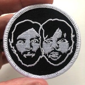 May include: A black and white embroidered patch featuring two faces, side by side, with white outlines. The patch has a white border.