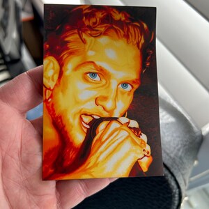 May include: A rectangular sticker featuring a portrait of a person with orange hair and blue eyes. The person is biting their hand. The artwork has a warm color palette with shades of orange, red, and brown.