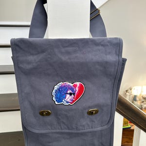 May include: A gray canvas messenger bag with a patch of a person's face with a red heart on it.