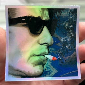 May include: Holographic sticker featuring a side profile of a person wearing sunglasses, smoking a cigarette. The image has a green and blue color scheme with a swirling background. The sticker has a silver border.