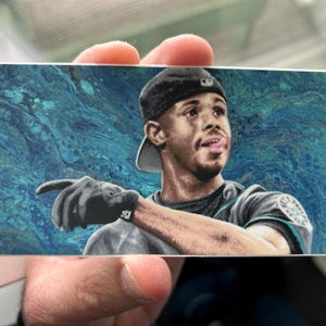 May include: A baseball player wearing a blue and white jersey with a black cap, pointing with his right hand. The background is a blue and green abstract design.