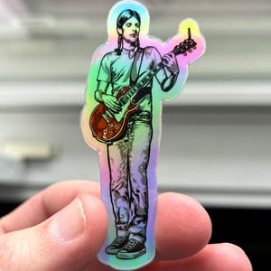May include: A holographic sticker featuring a person playing a red electric guitar. The person is wearing a white shirt and blue jeans. The sticker has a rainbow iridescent finish.