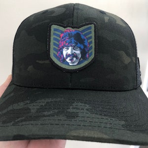 May include: A black and green camouflage trucker hat with a patch featuring a cartoon illustration of a man with long hair and a mustache. The patch has a blue, red, and white striped background.