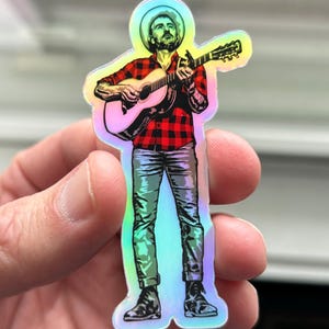 May include: A holographic sticker of a person wearing a cowboy hat and plaid shirt playing a guitar.