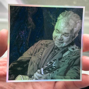 May include: A black and white image of a man playing a guitar. He is wearing a dark jacket with a floral pattern and has a serious expression on his face.