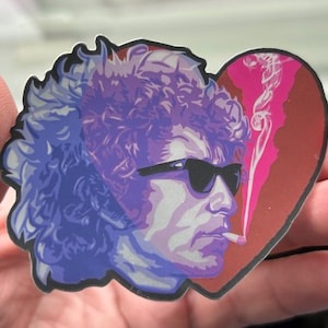 May include: A sticker of a man with curly hair wearing sunglasses and smoking a cigarette. The man is depicted inside a pink and red heart with a pink smoke trail.