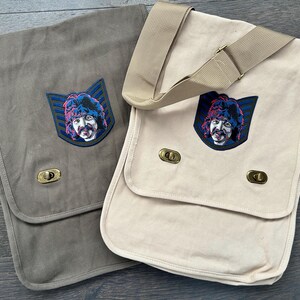 May include: Two canvas messenger bags in gray and cream. Each bag features a flap closure, a gold-colored clasp, and a shoulder strap. A colorful embroidered patch of a man's face is on the front of each bag.