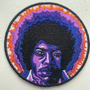 May include: Embroidered patch featuring a purple and blue portrait of a person with an afro hairstyle. The background is a circle with orange and purple flames.