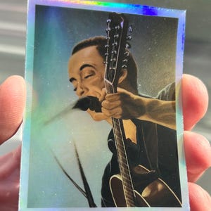 May include: A close-up of a man playing a guitar. He is wearing a black shirt and has a microphone in his mouth. The guitar is brown and has a silver headstock. The background is a light blue.