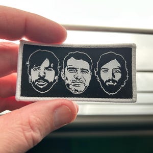 May include: A rectangular black and white patch featuring three stylized portraits of men with beards and various hairstyles. The patch has a white border and is held by a hand.