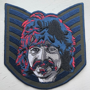 May include: An embroidered patch featuring a cartoon portrait of a man with long, curly hair, a mustache, and a beard. The patch is in a blue, red, and black color scheme and is set against a green background with blue stripes.