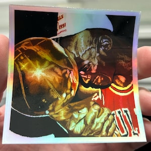 May include: A close-up of a basketball player holding a championship trophy. The player is wearing a red and white jersey with the number 23 on it. The trophy is gold and has the words "1991 Champions" on it.
