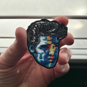 May include: Embroidered patch featuring a stylized face with black hair and a blue and yellow split design. The patch is held in a hand, showcasing the intricate details of the threadwork and the vibrant color palette.