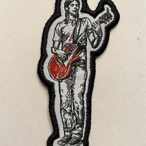 May include: An embroidered iron-on patch featuring a black and white illustration of a person playing a red electric guitar.