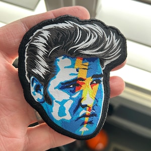 May include: An embroidered patch featuring a colorful portrait of Elvis Presley with a blue, yellow, and red lightning bolt design across his face.