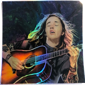 May include: A holographic sticker featuring a musician playing an acoustic guitar. The image shows a person with long hair and tattoos, strumming the guitar with their eyes closed. The guitar has a brown and orange finish.