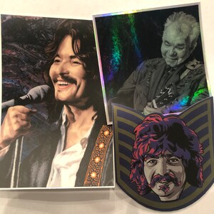 May include: Two colorful stickers featuring portraits of musicians. The first sticker shows a man with long dark hair and a microphone in his hand. The second sticker shows a man with short gray hair and a guitar in his hand. Both stickers have a holographic background.