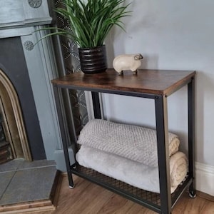 May include: A black metal and wood side table with two shelves. The top shelf is brown wood and the bottom shelf is a black metal wire grid. There are two white towels on the bottom shelf. A small white ceramic sheep figurine is on the top shelf.
