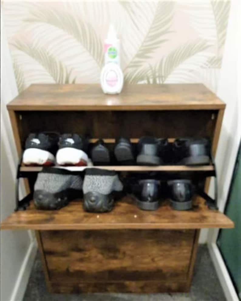 May include: A brown wood-grain shoe cabinet with a drop-down door featuring two internal shelves. The cabinet holds several pairs of shoes, including black sneakers and grey textured slip-ons. A small white bottle sits on the top surface.