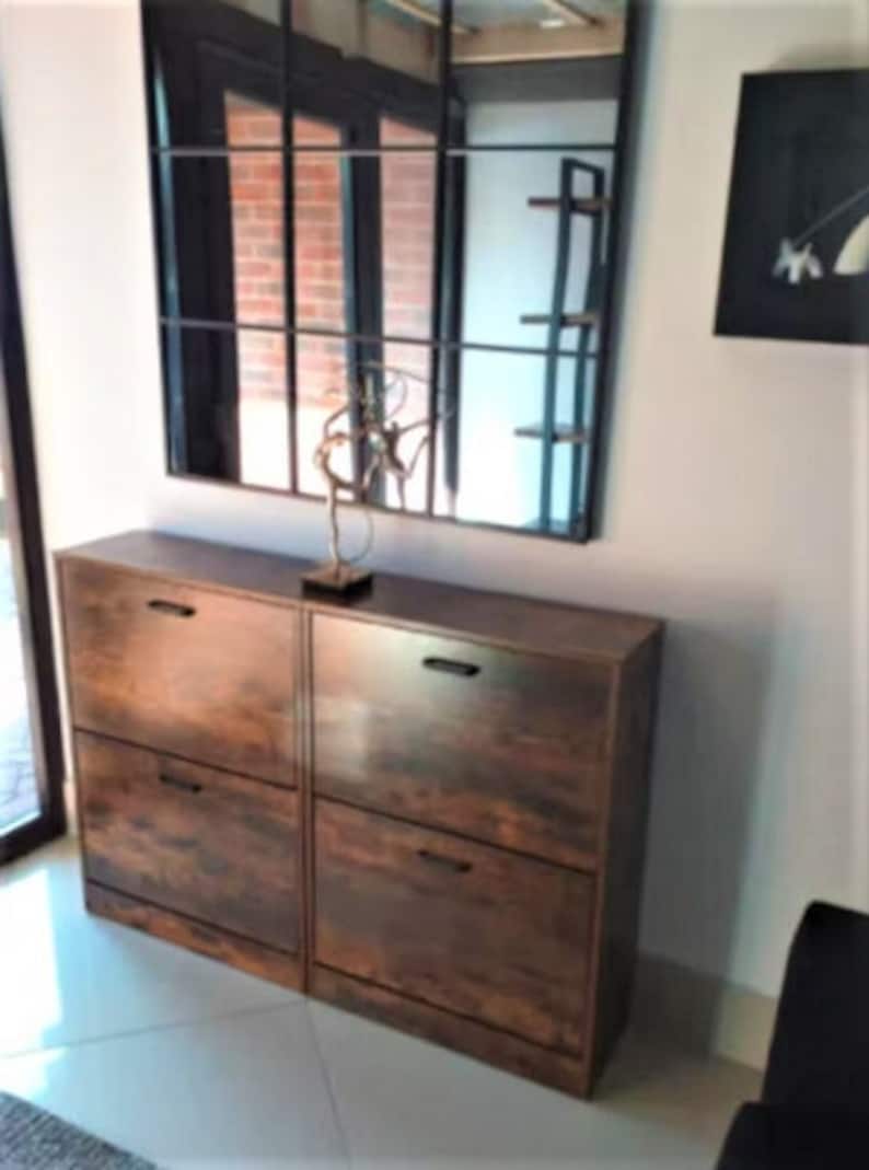 May include: A dark brown wood-grain shoe cabinet featuring four pull-down compartments with black horizontal handles. The cabinet is positioned beneath a large rectangular wall mirror with a black grid frame and a decorative metal sculpture.