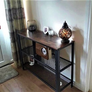 May include: A black metal console table with a brown wood top and two wire shelves. The table has a decorative lantern with a mosaic design, a small framed picture, and a radio on the top shelf.