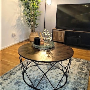 Pu&ograve; includere: Un tavolino da caff&egrave; rotondo in legno con una struttura in metallo nero. Il tavolo ha un design geometrico e si trova su un tappeto con un motivo blu chiaro e grigio.