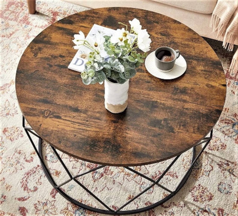 Pu&ograve; includere: Un tavolino da caff&egrave; rotondo in legno con una struttura in metallo nero. Il tavolo ha una finitura in legno invecchiato e un design geometrico sul telaio. Un mazzo di fiori bianchi in un vaso bianco si trova sul tavolo, insieme a una tazza di caff&egrave; e un piattino.