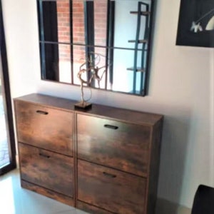May include: A dark brown wood-grain shoe cabinet featuring four pull-down compartments with black horizontal handles. The cabinet is positioned beneath a large rectangular wall mirror with a black grid frame and a decorative metal sculpture.