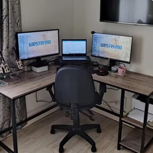 May include: A corner desk with a black metal frame and a wood grain laminate top. The desk has a monitor on each side and a laptop in the middle. A black office chair sits in front of the desk.