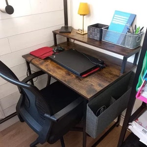 May include: A home office desk with a dark brown wood-grain finish, black metal frame, and a black office chair. The desk has a top shelf with a yellow lamp, a woven basket, and a wire basket with pens. A laptop, red mousepad, and a black mouse are on the desk.