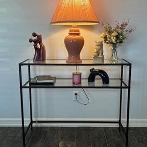 May include: A black metal console table with two glass shelves. The top shelf holds a lamp with a peach-coloured shade, a sculpture, and a vase of flowers. The bottom shelf has a book, a candle, and a dark blue arch sculpture.