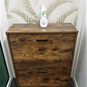 May include: A rectangular wooden shoe cabinet with a dark brown wood grain finish, featuring two pull-down drawers with black horizontal handles. A white plastic bottle with a pump dispenser sits on the top surface of the cabinet.