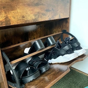 May include: A wooden shoe rack with two shelves. The shelves are filled with black sandals and sneakers. The shoe rack has a brown wood finish.