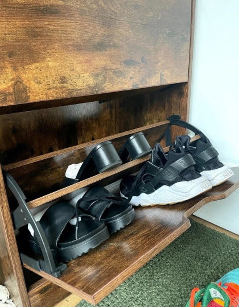 May include: A wooden shoe rack with two shelves. The shelves are filled with black sandals and sneakers. The shoe rack has a brown wood finish.