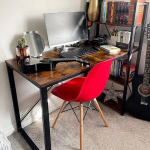May include: A brown wooden desk with black metal legs and a red chair. The desk has a computer monitor, keyboard, and a guitar on top. There is a bookshelf behind the desk with books and a decorative vase.