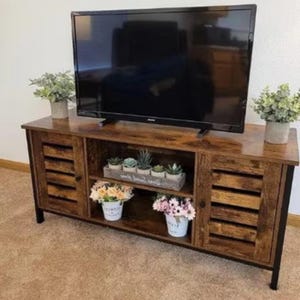 May include: A rustic brown TV stand with two doors and a shelf. The doors have a slatted design and the shelf is open. There are plants and a sign on the shelf. The stand has black metal legs.