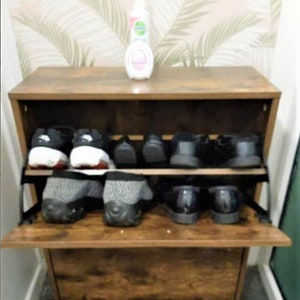 May include: A brown wood-grain shoe cabinet with a drop-down door featuring two internal shelves. The cabinet holds several pairs of shoes, including black sneakers and grey textured slip-ons. A small white bottle sits on the top surface.
