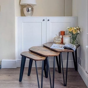 May include: Three wooden nesting tables with black metal hairpin legs. The tables are arranged in a staggered formation, with the largest table at the back and the smallest table at the front. The tables are made of a dark wood with a distressed finish.