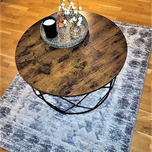 Pu&ograve; includere: Un tavolino da caff&egrave; rotondo in legno con una struttura in metallo nero si trova su un tappeto a motivi grigi e bianchi. Un vassoio decorativo con una candela, un piccolo vaso con fiori bianchi e un vaso di vetro con fiori bianchi si trova sul tavolo.