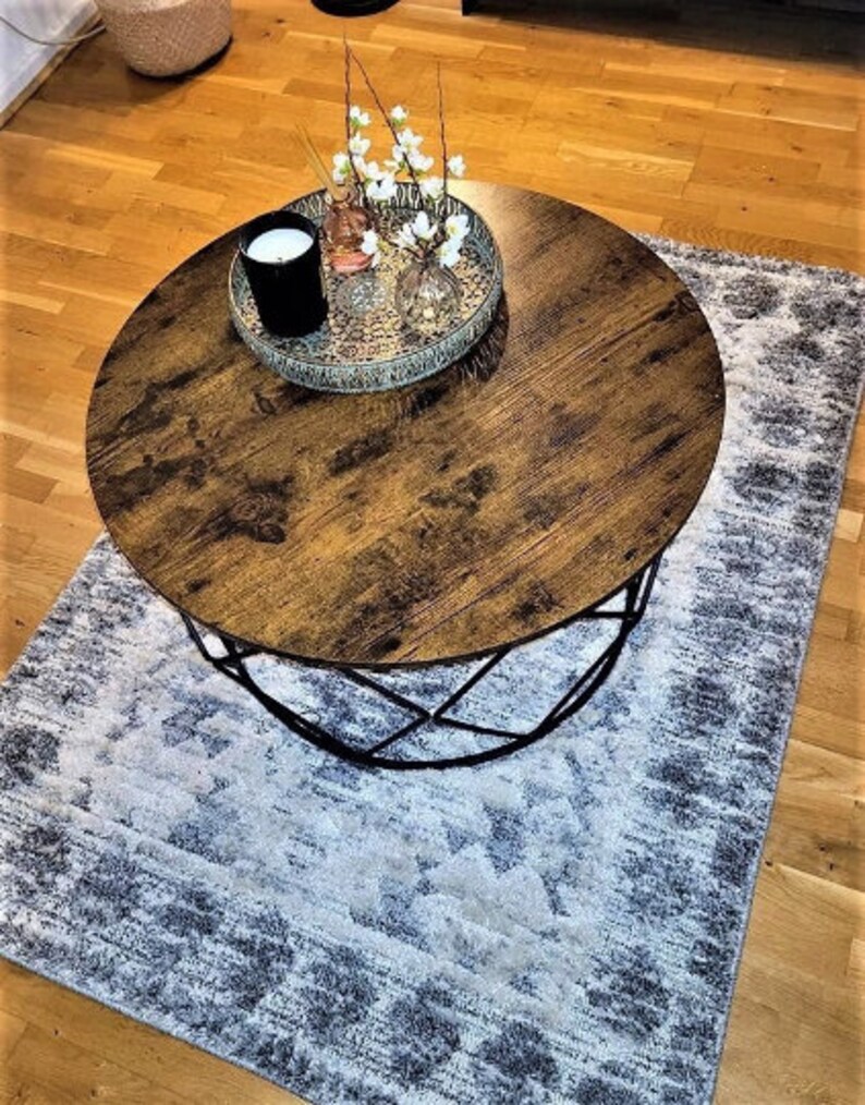 Pu&ograve; includere: Un tavolino da caff&egrave; rotondo in legno con una struttura in metallo nero si trova su un tappeto a motivi grigi e bianchi. Un vassoio decorativo con una candela, un piccolo vaso con fiori bianchi e un vaso di vetro con fiori bianchi si trova sul tavolo.