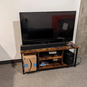 May include: A dark brown wooden TV stand with black metal legs and shelves. The stand has a flat top surface with a black soundbar and a flat screen TV on top. There is a cardboard box on the bottom shelf and a black speaker on the right side.