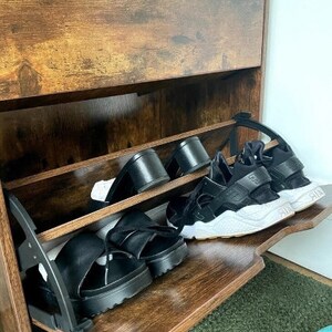May include: A wooden shoe rack with two shelves. The shelves are filled with black sandals and sneakers. The shoe rack has a brown wood finish.