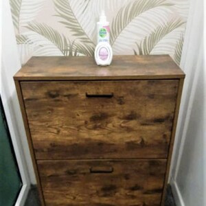May include: A rectangular wooden shoe cabinet with a dark brown wood grain finish, featuring two pull-down drawers with black horizontal handles. A white plastic bottle with a pump dispenser sits on the top surface of the cabinet.