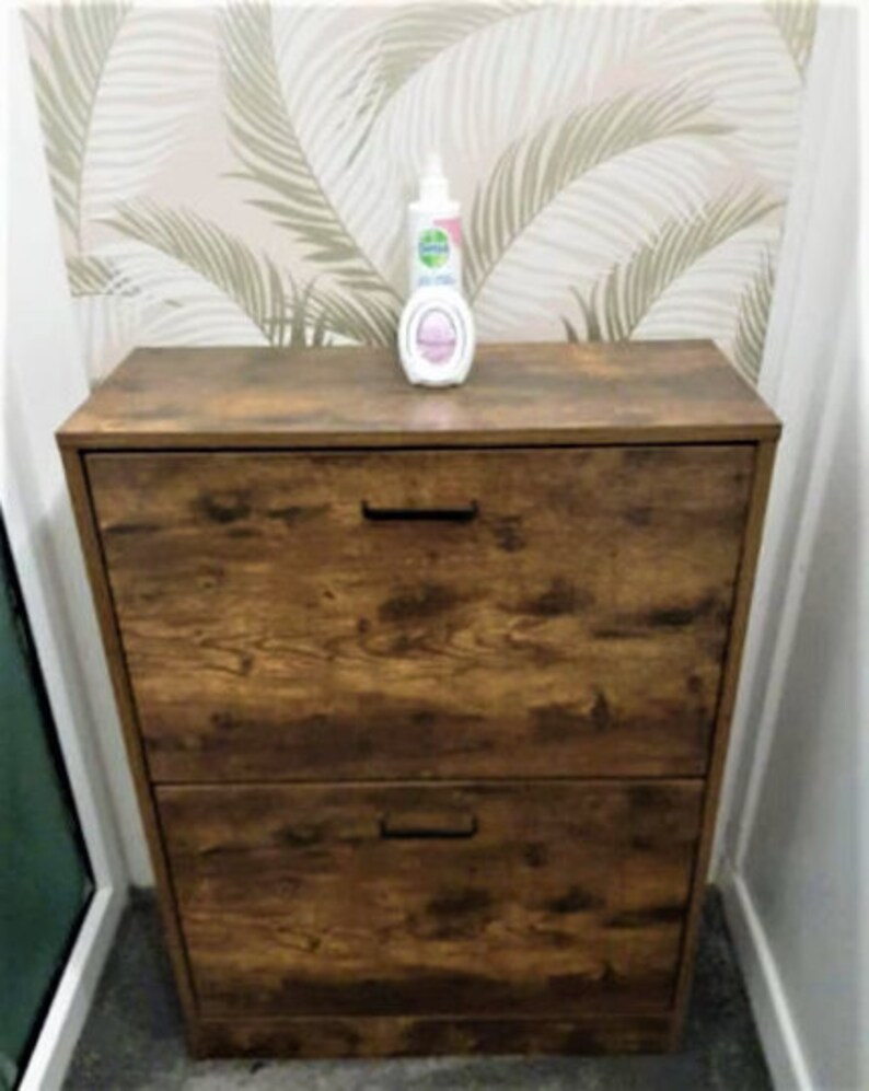 May include: A rectangular wooden shoe cabinet with a dark brown wood grain finish, featuring two pull-down drawers with black horizontal handles. A white plastic bottle with a pump dispenser sits on the top surface of the cabinet.