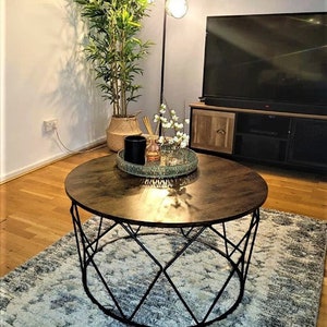 Pu&ograve; includere: Un tavolino da caff&egrave; rotondo in legno con una struttura in metallo nero. Il tavolo ha un design geometrico e si trova su un tappeto con un motivo blu chiaro e grigio.