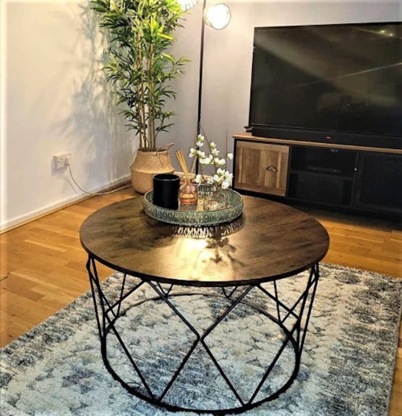 Pu&ograve; includere: Un tavolino da caff&egrave; rotondo in legno con una struttura in metallo nero. Il tavolo ha un design geometrico e si trova su un tappeto con un motivo blu chiaro e grigio.