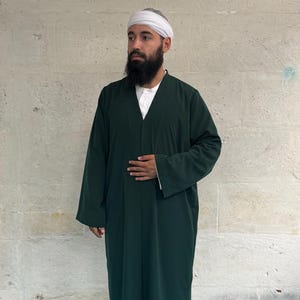 May include: A dark green long-sleeved robe with a V-neckline worn over a white collared shirt and light-colored trousers. The ensemble includes a white head wrap and black socks, presenting a layered look with a solid dark green outer garment.